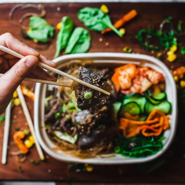 Chopsticks holding a piece of Matko's Korean BBQ beef over a takeout container with kimchi and noodles.