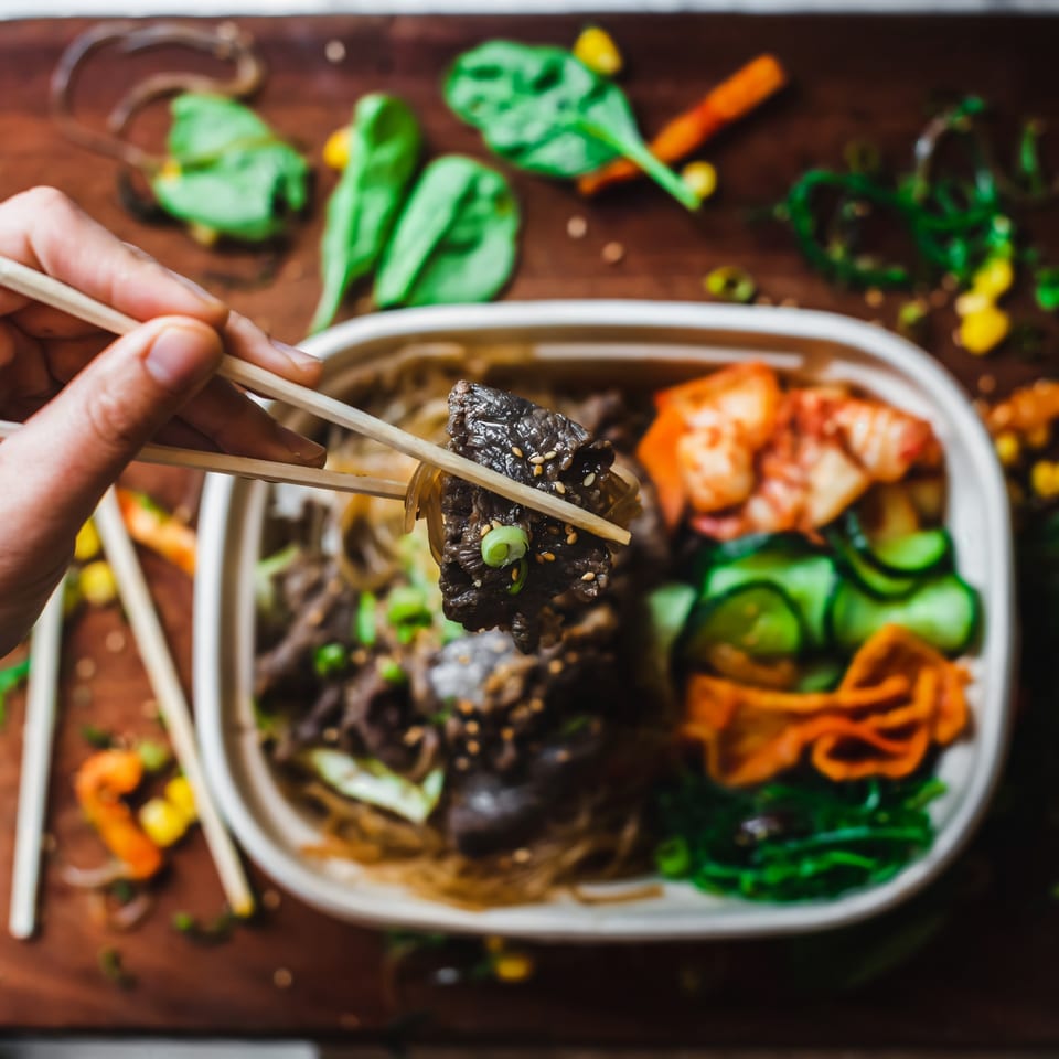 Chopsticks holding a piece of Matko's Korean BBQ beef over a takeout container with kimchi and noodles.