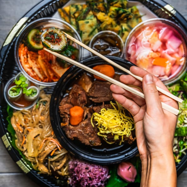 A hand holds chopsticks with a cucumber kimchi roll over a Korean bento box with various dishes.