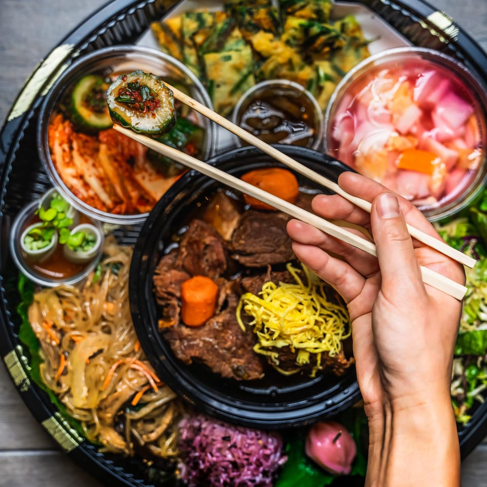 A hand holds chopsticks with a cucumber kimchi roll over a Korean bento box with various dishes.